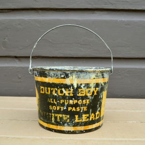 May include: An antique Dutch Boy paint bucket with a metal handle. The black bucket has yellow stripes and text that reads "DUTCH BOY ALL-PURPOSE SOFT PASTE WHITE LEAD". The bucket shows signs of wear and age.