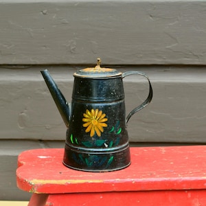May include: Black metal teapot with a yellow daisy design. The teapot has a long spout, a handle, and a lid. The teapot is sitting on a red wooden stool. The background is a brown wooden wall.