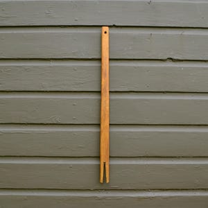 May include: A long, slender, wooden weaving shuttle with a forked end and a hole at the top. The shuttle is a warm, natural wood tone, set against a gray-green wooden background. This tool is used in weaving.