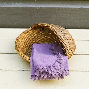 May include: A woven wicker basket with a purple fringed throw blanket. The basket is brown and has a rounded shape. The blanket is a solid lavender color and is partially draped over the basket.