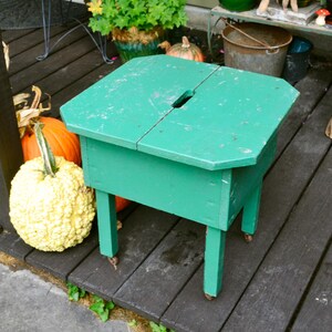 K&ouml;nnte beinhalten: Ein gr&uuml;ner Holzhocker mit einer klappbaren Oberseite. Der Hocker hat ein kleines Loch oben und steht auf einem Holzdeck. Im Hintergrund sind K&uuml;rbisse zu sehen.