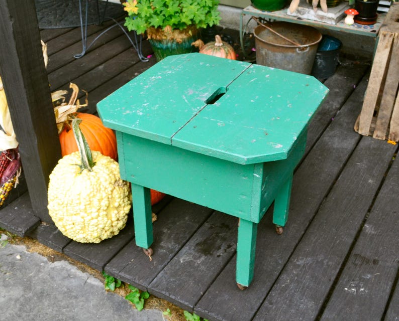 K&ouml;nnte beinhalten: Ein gr&uuml;ner Holzhocker mit einer klappbaren Oberseite. Der Hocker hat ein kleines Loch oben und steht auf einem Holzdeck. Im Hintergrund sind K&uuml;rbisse zu sehen.