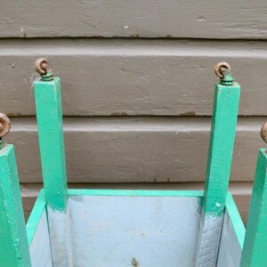 K&ouml;nnte beinhalten: Ein gr&uuml;ner Holz-Pflanzkasten mit vier Metallr&auml;dern. Der Pflanzkasten hat einen hellblauen Innenraum und ist bereit, Pflanzen aufzunehmen.