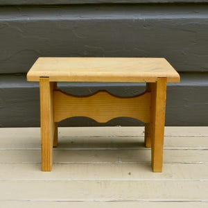 May include: A small, light-colored wooden stool with a rectangular top and four straight legs. A horizontal support piece connects the legs, featuring a decorative scalloped cutout along its bottom edge. The wood grain is visible throughout the surface.