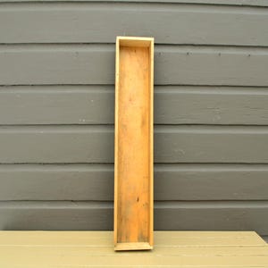 May include: A long, rectangular wooden box with a natural finish. The box is open at the top and has a simple, minimalist design. The box is sitting on a light-colored wooden surface against a gray background.