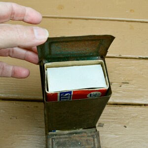 May include: A vintage metal matchbox with a worn, rusty exterior. The matchbox is open, revealing a full box of matches with a red, white and blue label that reads "Strike's".