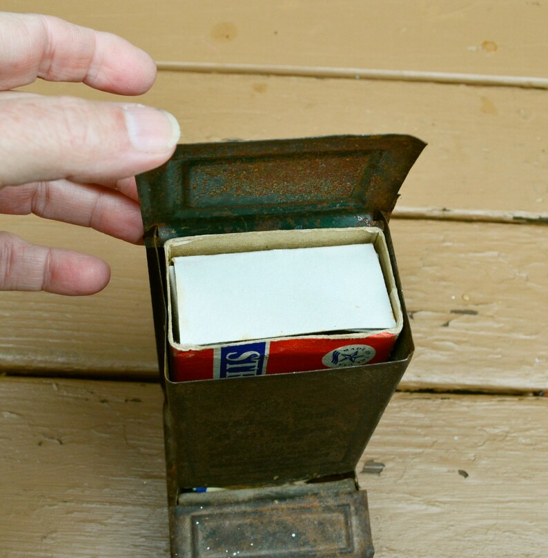 May include: A vintage metal matchbox with a worn, rusty exterior. The matchbox is open, revealing a full box of matches with a red, white and blue label that reads "Strike's".