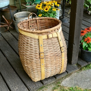 May include: A large, woven basket with a wide, open top and a rounded base. The basket is made of light brown wicker and has two tan straps with buckles. The basket is sitting on a wooden deck.