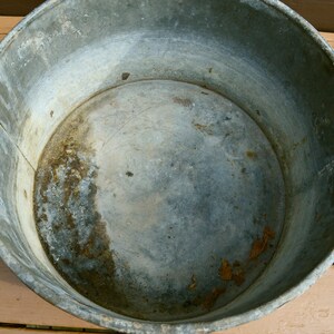 May include: A large, round, galvanised metal tub with a slightly dented bottom. The tub is empty and has a worn, aged appearance.