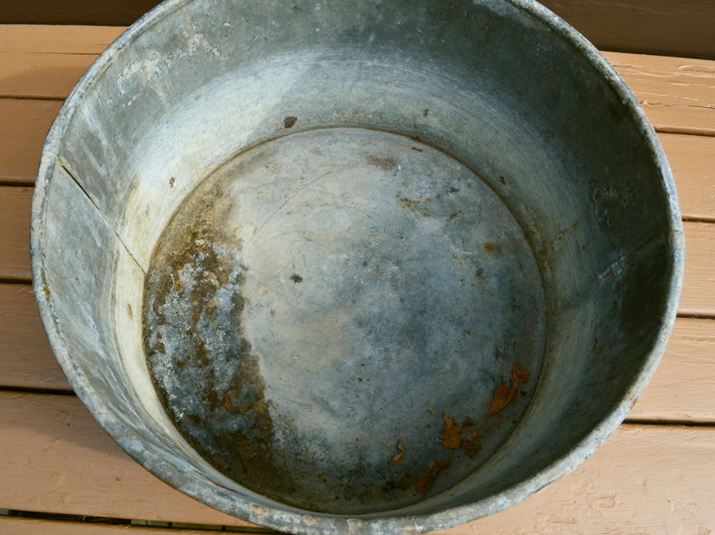 May include: A large, round, galvanised metal tub with a slightly dented bottom. The tub is empty and has a worn, aged appearance.