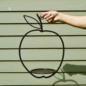May include: A black metal wire frame shaped like an apple with a single leaf at the top. The structure features a circular base platform at the bottom, designed to hold an object. The entire piece is constructed from thin, dark metal rods.