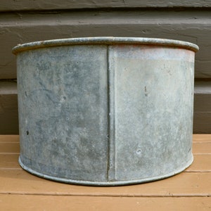 May include: A large, round, galvanised metal planter with a slightly distressed finish. The planter is empty and has a slightly rounded bottom.