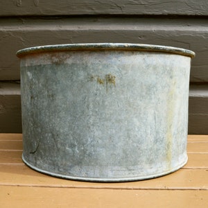 May include: A large, round, galvanised metal planter with a slightly dented rim. The planter is weathered and has a rustic appearance.