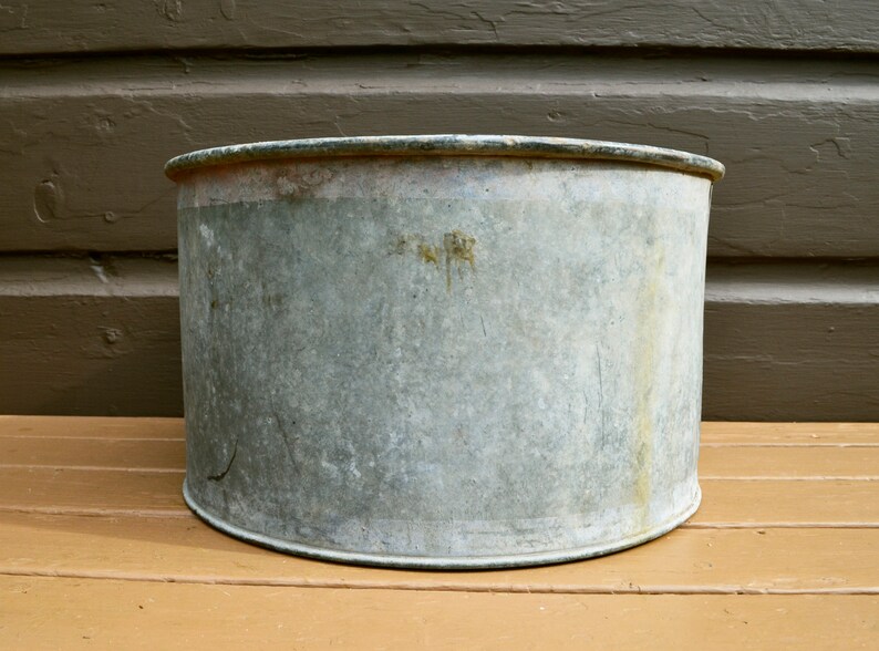 May include: A large, round, galvanised metal planter with a slightly dented rim. The planter is weathered and has a rustic appearance.