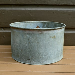 May include: A weathered, galvanised metal planter with a round, wide opening. The planter is empty and has a light grey colour.