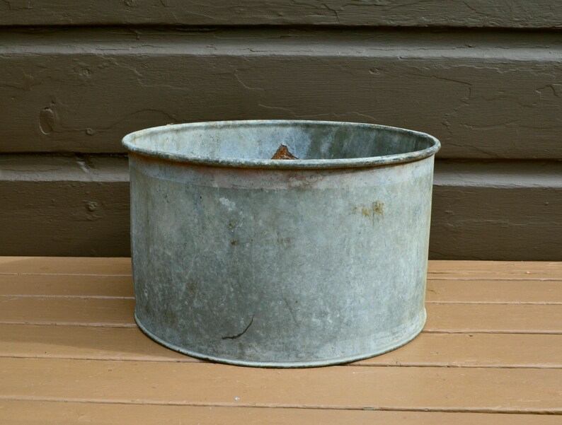May include: A weathered, galvanised metal planter with a round, wide opening. The planter is empty and has a light grey colour.