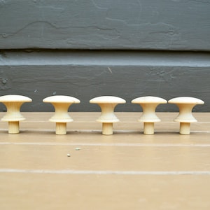 May include: Six wooden knobs in a row on a light brown surface. The knobs are a light beige color and have a rounded top and a short stem.
