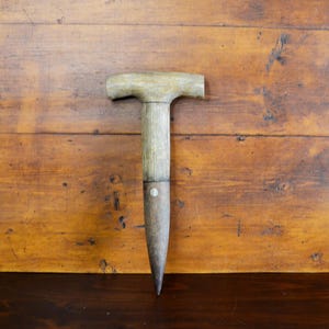 May include: A vintage, T-shaped garden tool with a wooden handle and a metal blade. The handle is light-colored, and the blade is dark, with a pointed end. The tool is set against a wooden background.