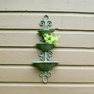 May include: A vertical wall-mounted planter featuring three tiered, fluted, bowl-shaped containers connected by ornate, scrolled metalwork. The structure has a weathered green finish and contains a small cluster of bright green artificial foliage.
