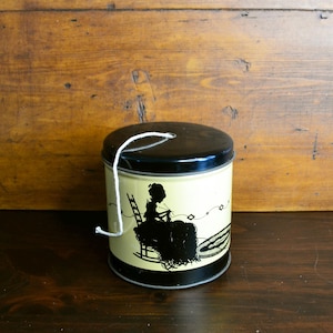 May include: A vintage metal tin with a black lid and base, and a cream-colored body. The tin features a black silhouette of a person knitting on a rocking chair. A white string is attached to the top.