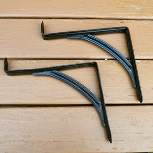 May include: Two black metal shelf brackets with a curved design. The brackets are on a light brown wooden surface.