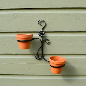 May include: A black wrought iron wall-mounted planter with two terracotta pots. The pots are held in place by black metal rings. The planter has a decorative floral element at the top and is mounted on a light green wooden wall.