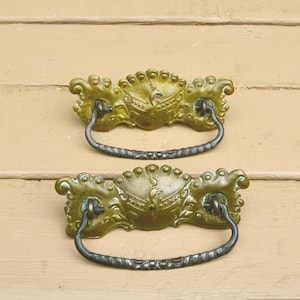 May include: Two antique brass drawer pulls with ornate detailing. The pulls have a dark, curved handle and a decorative backplate with a crest design. The brass has a greenish patina, suggesting age and use. These pulls are likely for a cabinet or dresser.