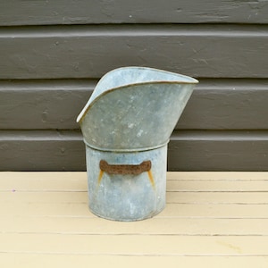 May include: A vintage galvanized metal coal scuttle with a curved top and a cylindrical base. The scuttle has a handle with rust-colored accents. The background features a wooden wall and a light-colored wooden surface.