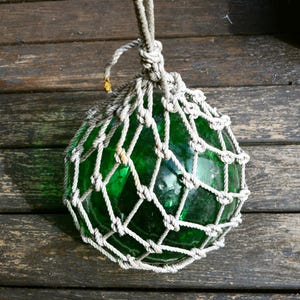 May include: A round, green glass float encased in a white knotted net, suspended by a thick rope. The float is a vibrant green color and appears to be made of glass. The net is tightly woven, and the rope is knotted at the top.