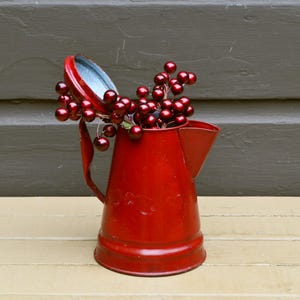 May include: A bright red metal pitcher with a flared base and a side handle, featuring a hinged lid that is propped open. Inside the pitcher are several stems of shiny, dark red decorative berries on thin wire branches, creating a festive display.