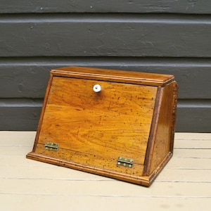 May include: A wooden bread box with a slanted front door featuring a speckled brown finish, a small white ceramic knob, and two brass-colored metal hinges at the base. The box has a rectangular base and a sloped top surface.
