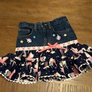 May include: A denim skirt with a unicorn print fabric ruffle and a denim top with embroidered unicorns and stars.