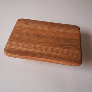 May include: A rectangular wooden cutting board with rounded corners. The board is made of light brown wood with visible grain patterns. The board is angled slightly, giving a sense of depth.