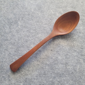 May include: A wooden spoon with a long handle and a rounded bowl. The spoon is made of a reddish-brown wood and has a smooth, polished finish. The spoon is set against a gray background.