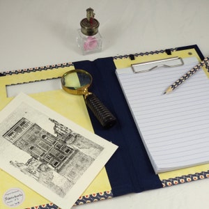 May include: A blue and yellow clipboard with a white lined notepad, a pencil, and a magnifying glass. The clipboard has a decorative pattern and a small, clear glass bottle with a pink object inside.