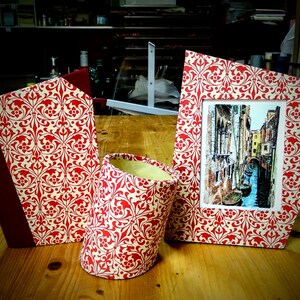 Può includere: Tre oggetti decorativi con un design a motivi rossi e bianchi. Un libro, una cornice per foto e un contenitore cilindrico.