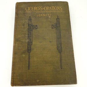 May include: A vintage book titled "CICERO'S ORATIONS" by Jennett. The cover is a textured olive green with two black line drawings of a pole with a flag. The book has a worn appearance.