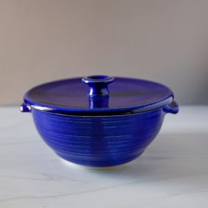 May include: A blue ceramic casserole dish with a lid. The dish has a slightly rounded shape and a smooth, glossy finish.