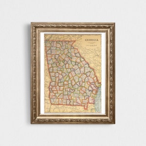 May include: A vintage map of Georgia, framed in a gold ornate frame. The map shows the state's counties and major cities. The map is printed on aged paper with a cream background and brown lines.