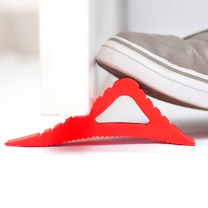 Puede incluir: Un tope de puerta de plástico rojo en forma de triángulo con bordes redondeados está siendo mantenido en su lugar por el pie de una persona que lleva un zapato gris y azul.