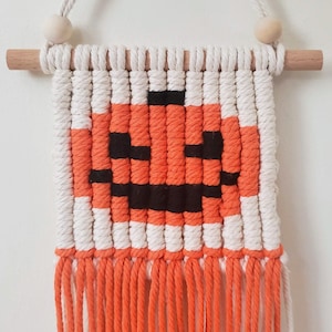 May include: A macrame wall hanging with a pumpkin design. The pumpkin is made with orange and black yarn and has a white background. The wall hanging has a fringe of orange yarn.