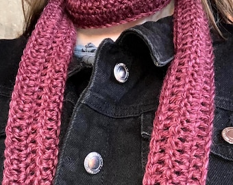 May include: A burgundy crochet scarf is draped over a black denim jacket. The scarf is made of a thick yarn, with a textured, woven appearance. The jacket has silver buttons.