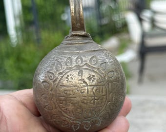 May include: Antique bronze bell with a loop handle. The bell has a round body with etched floral and text details. The bell is held in a hand, with a blurred outdoor background.