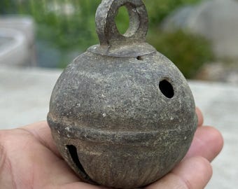 May include: A weathered, round metal bell with a loop at the top for hanging. The bell has a dark, textured surface with two holes and a slit. The bell is held in a hand, suggesting its size and portability. This item is likely a decorative or functional piece.