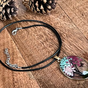 Puede incluir: Un collar de cord&oacute;n negro con un cierre plateado y un colgante redondo con flores prensadas y una mariposa dentro de un &oacute;valo de resina transparente.