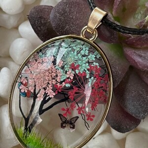 Puede incluir: Un collar con un colgante ovalado de oro y una c&uacute;pula de vidrio transparente que contiene flores prensadas y una mariposa. Las flores son rosas, rojas y verdes.