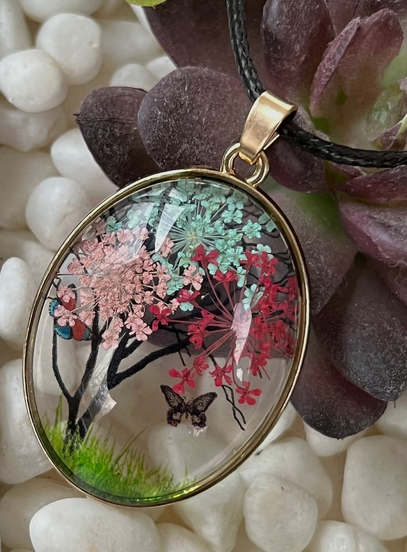 Puede incluir: Un collar con un colgante ovalado de oro y una c&uacute;pula de vidrio transparente que contiene flores prensadas y una mariposa. Las flores son rosas, rojas y verdes.