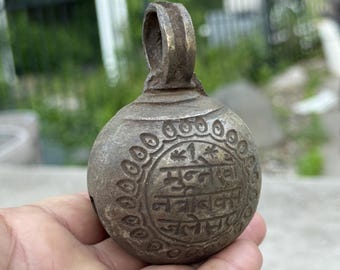 May include: Antique bronze bell with a loop handle. The bell has an engraved design around the bottom with text in a circular pattern. The bell is held in a hand against a blurred green background.