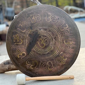 May include: A large, round, bronze gong with a wooden mallet. The gong is decorated with a repeating pattern of the Om symbol in gold.