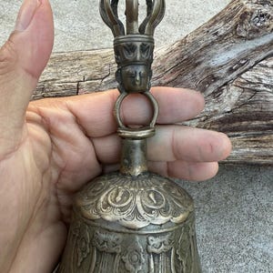 May include: A silver bell with intricate carvings and a crown-shaped handle. The bell has a detailed design of a face on the handle and floral patterns on the body.